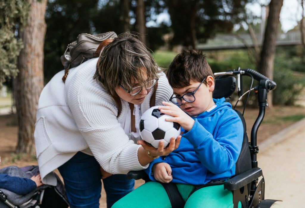 Barn med funktionshinder i rullstol, håller i en boll tillsammans med kvinna/mamma.
